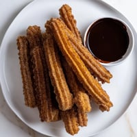Freshly fried churros coated in cinnamon sugar are arranged beside a cup of rich chocolate dipping sauce. 