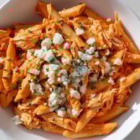 A close-up of Buffalo Chicken Pasta in a white bowl, featuring penne pasta coated in creamy orange sauce with shredded chicken, topped with crumbled blue cheese and fresh chives.