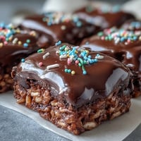 A close-up of Chocolate Covered Rice Krispy Treats, with glossy melted chocolate and vibrant rainbow sprinkles on top.